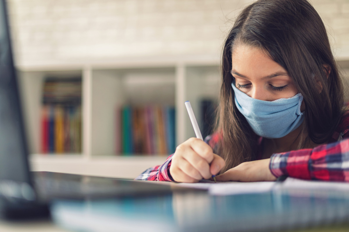 A child wearing a mask works on an assignment in front of a laptop