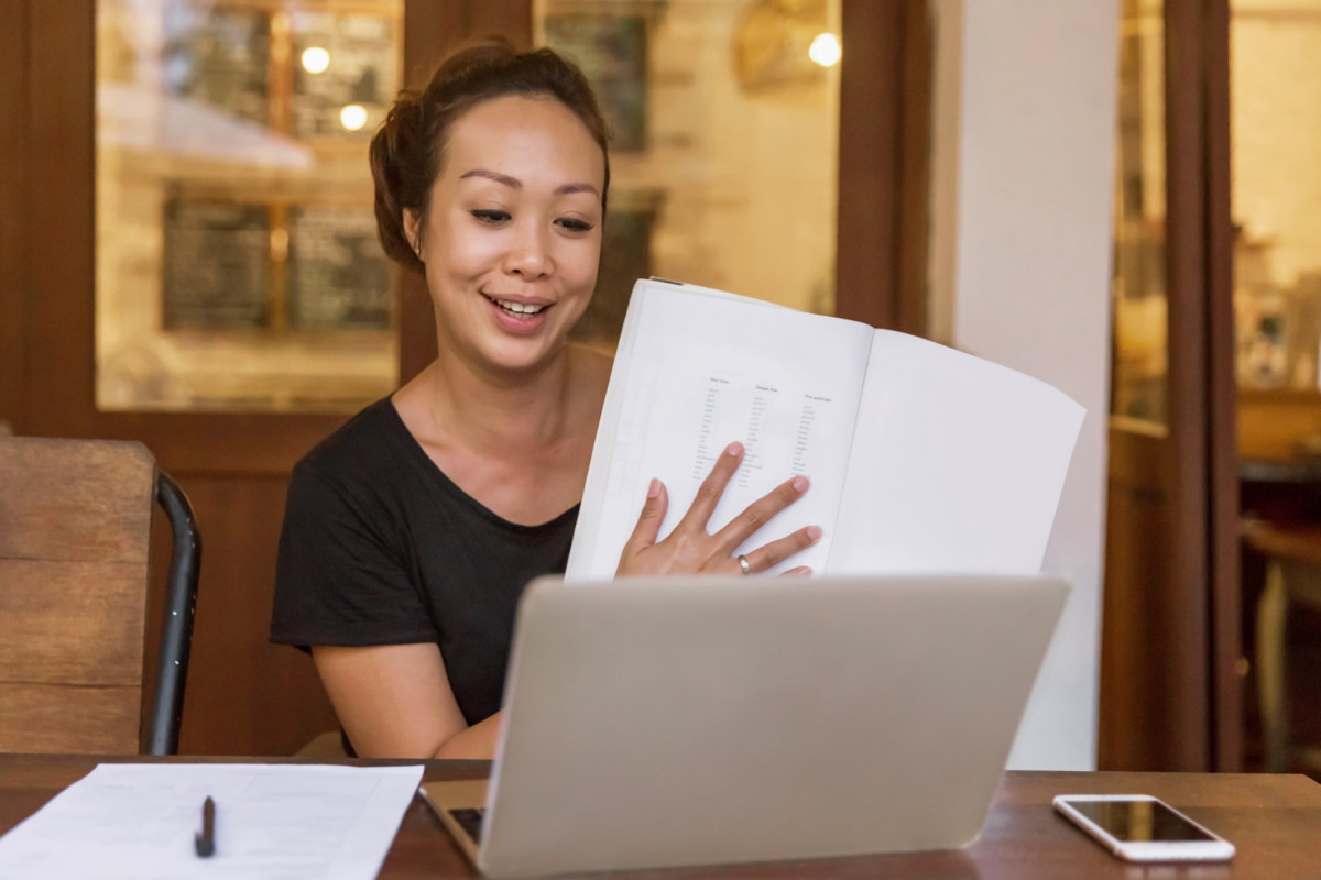 A teacher holds papers in front of a laptop screen