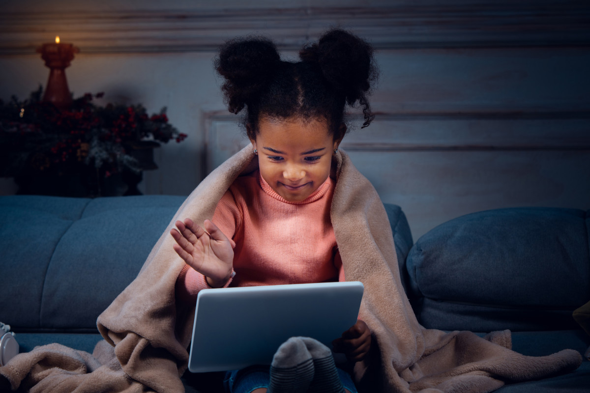 A student sitting on the couch wearing a blanket and waving at a laptop screen