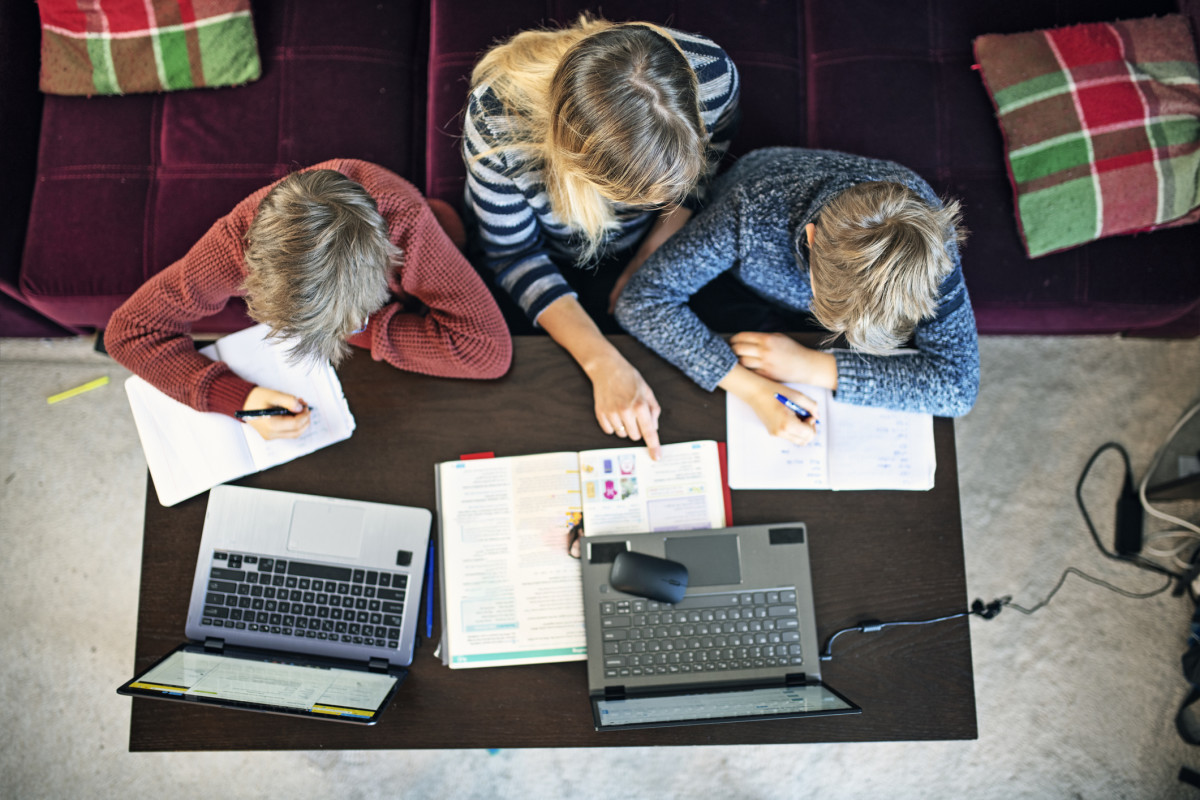 A caregiver instructs two children in the front room with their laptops in front of them