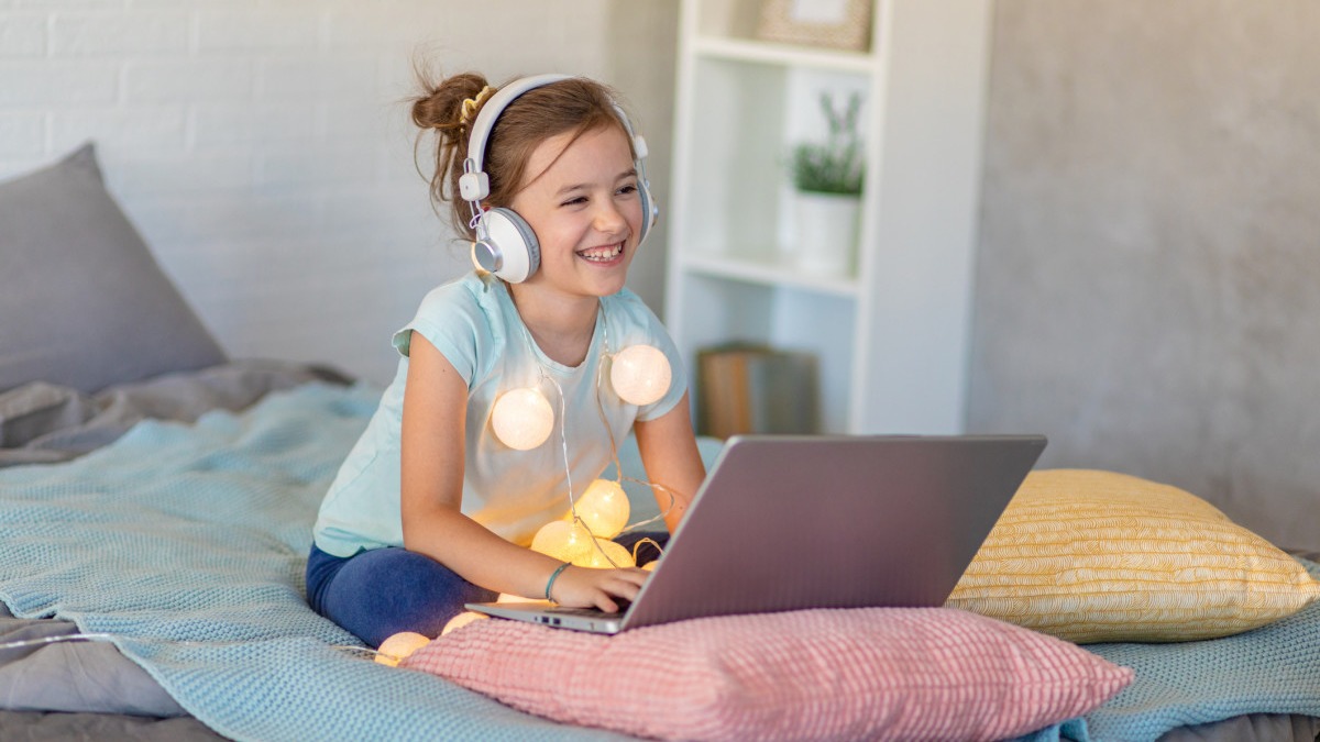 A girl wearing headphones sits with her laptop on her bed with a string of lights hanging from her neck