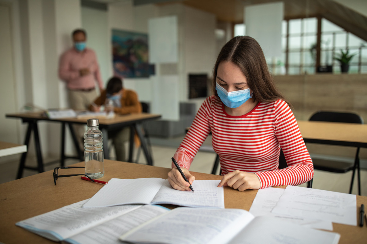 A teacher wearing a mask looks on at two students wearing masks working on class work