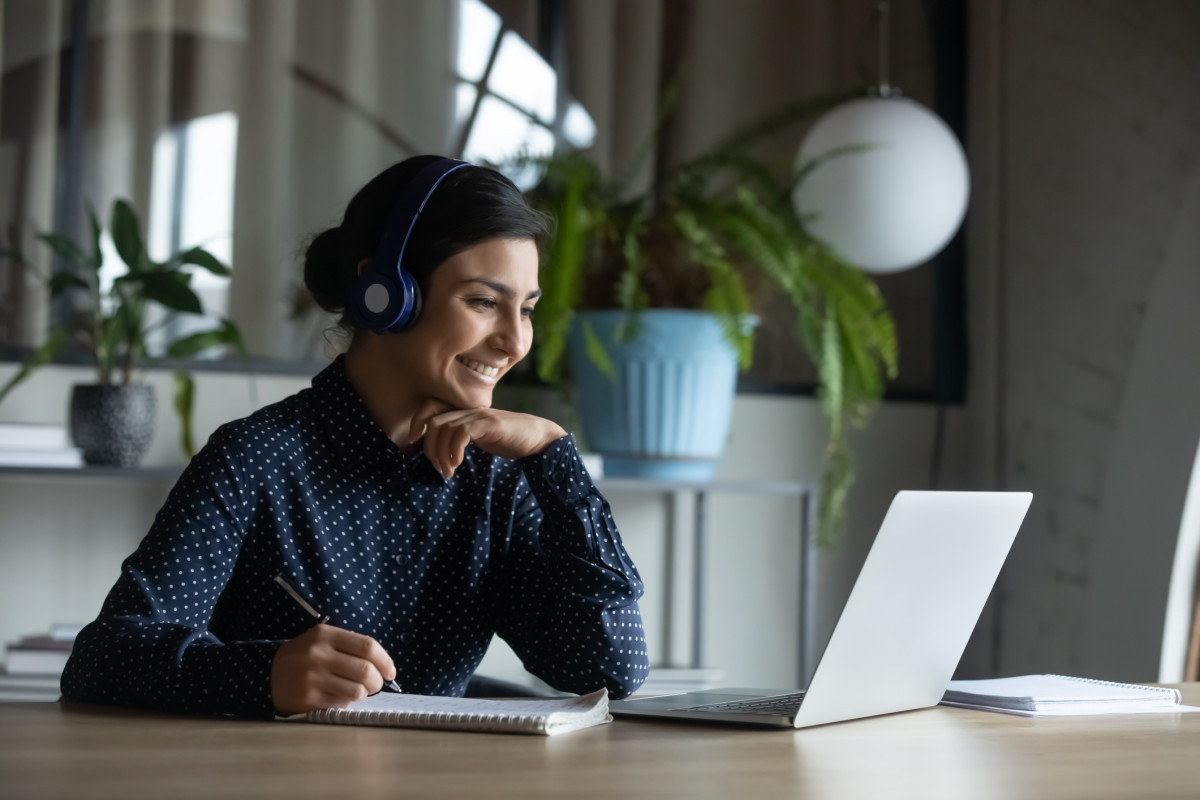 A teacher works on a laptop at home, writing notes in a notebook