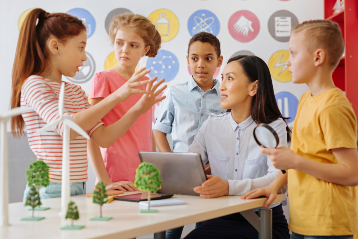 Photo of students explaining a concept to a teacher sitting at a table with a model of a wind turbine on the table