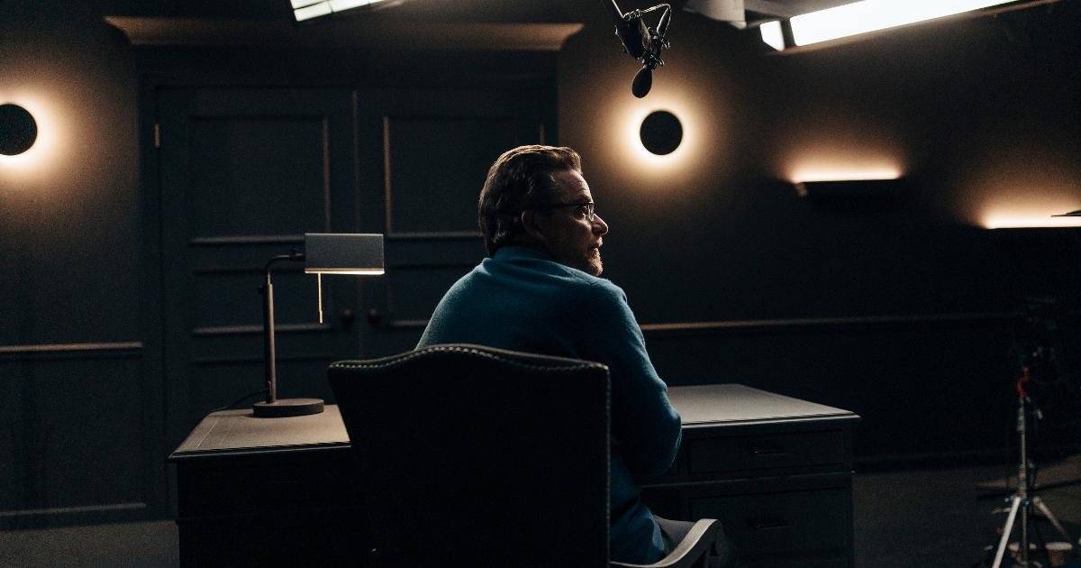 Man sitting in a dark room with golden lights on the wall and a microphone at his desk, he is looking off to the right