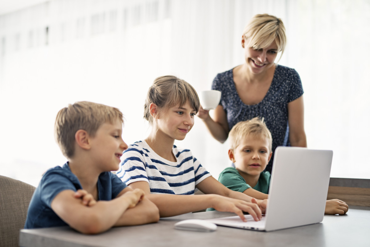 Three children sit together at a table on a laptop while a caregiver looks over their shoulders