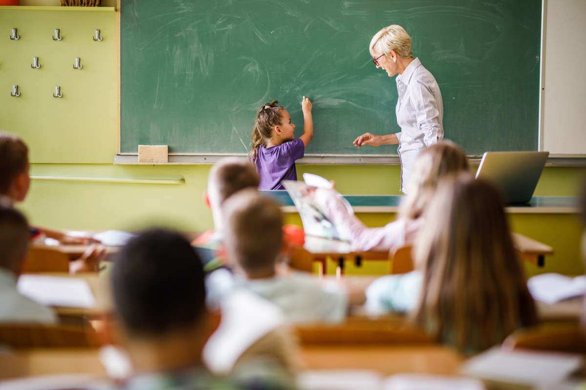 A young student writes on a chalk board at the front of the class while a teacher looks on