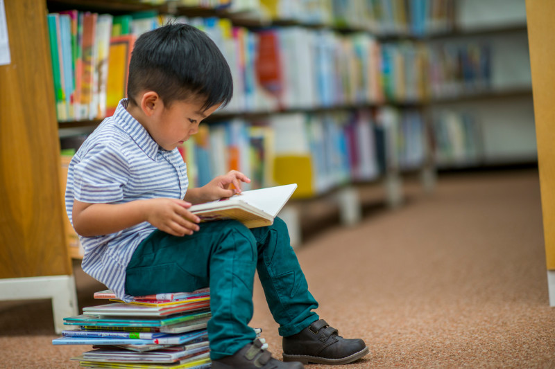 An image of a young child sitting on a stack of books in the library, reading