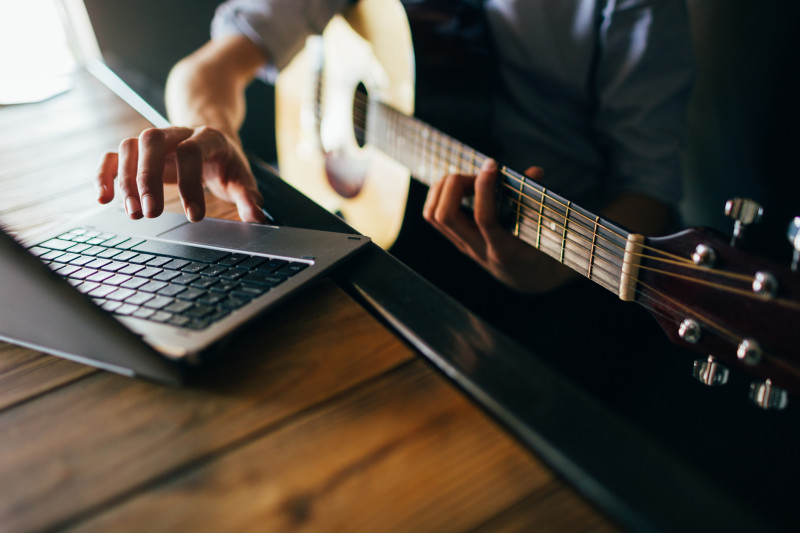 A person holds a guitar while typing on a laptop