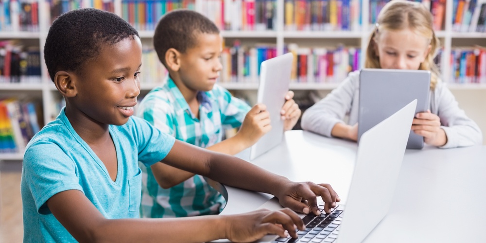 Three students sit at a table in the library. One uses a laptop, and the other two use tablets.