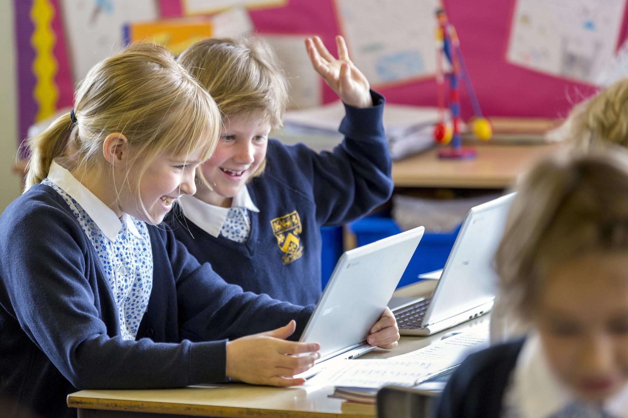 Two students look at their laptops with a hand raised