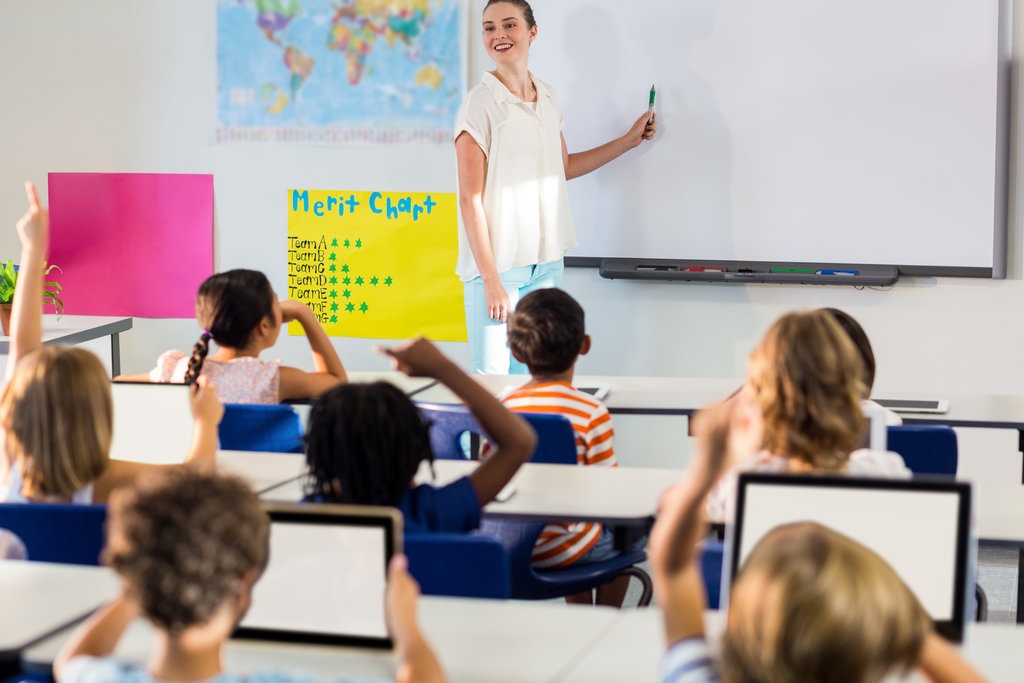 An image of a teacher pointing to a whiteboard at th front of a classroom with a student raising his hande