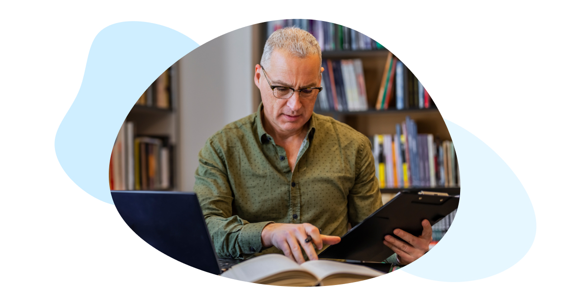 A teacher looks at a clipboard and book with his laptop open