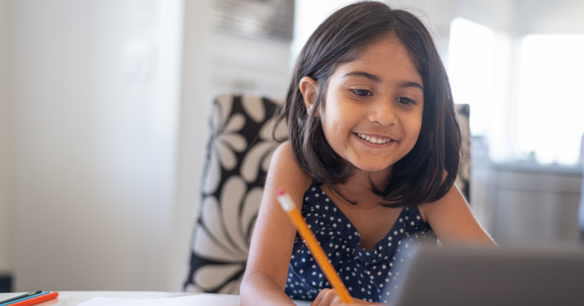 A young student smiles while looking at a laptop screen and writing down an answer